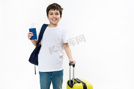 身穿蓝色牛仔裤和休闲 T 恤、背包、旅行袋和登机牌的快乐青春期男孩游客，在镜头前微笑