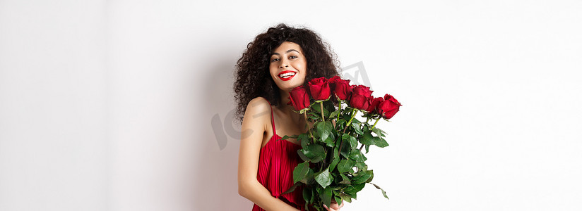 一头卷发、穿着时髦的浪漫女士，手捧红玫瑰花束，微笑着，站在白色背景上开心地站着