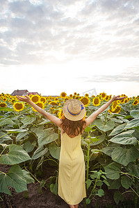 一个身穿黄色连衣裙、头戴草帽的女孩，双手高举，背负着手捧着一束向日葵站在一大片向日葵花田上。
