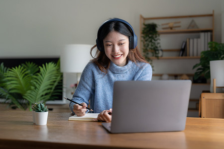 亚洲女商人摄影照片_微笑的女学生戴耳机与老师在线学习，快乐的亚洲年轻女性学习语言听讲座看网络研讨会写笔记看坐在咖啡馆里的笔记本电脑，远程教育