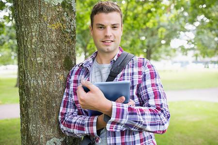 微笑的学生靠在树上拿着他的数字平板电脑