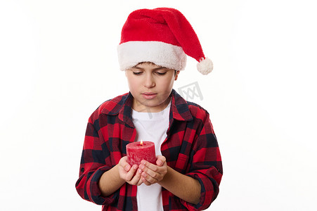 戴着圣诞帽的迷人白人男孩，拿着一根点燃的红蜡烛，为圣诞节许愿，在白色背景中与世隔绝