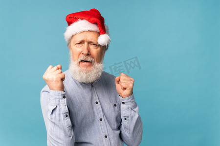 圣诞老人胡子帽子摄影照片_快乐自信、酷酷、留着胡子的圣诞老人赢家举起拳头，庆祝蓝色背景下的胜利和成功，并留有复制空间
