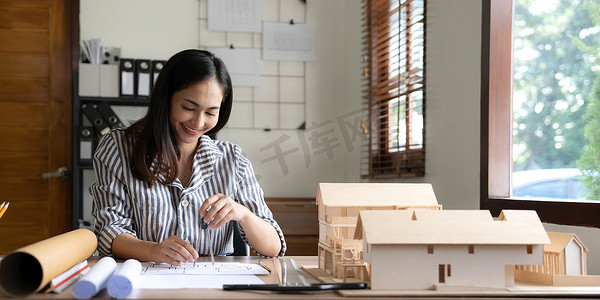 房地产意境摄影照片_建筑、建筑、施工和房地产概念-快乐微笑的女建筑师，在家庭办公室工作的桌子上有蓝图、尺子和生活房屋模型。