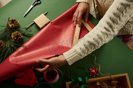 顶视图女人使用尺子，计算包装圣诞礼物所需的包装纸数量，绿色背景。