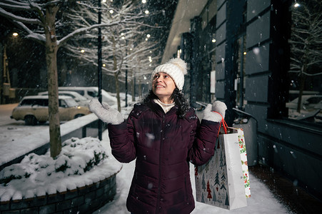 带着购物袋的令人愉快的女人，沿着一条被假日花环照亮的雪街散步时，抓着雪花
