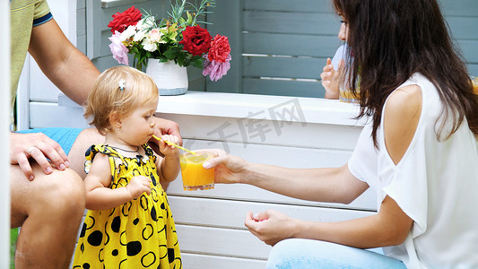 鲜榨果汁广告摄影照片_夏天，在花园里，父母带着年幼的孩子玩耍，一个女孩和一个男孩，在咖啡馆，在儿童游乐场，用鲜榨果汁招待孩子，喝果汁。