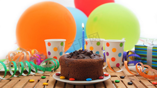 巧克力生日蛋糕，蓝色蜡烛在质朴的木桌上燃烧，背景是彩色气球、礼物、塑料杯和糖果，背景是白墙
