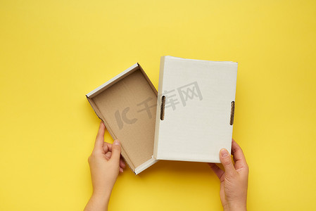 纸盒牛皮纸包装摄影照片_两只女手在黄色背景上拿着一个打开的空白纸盒