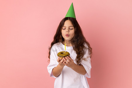 头戴绿色派对锥的可爱迷人小女孩拿着蛋糕，吹蜡烛，许愿。
