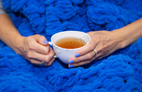 一个女孩裹着一条大的蓝色美利奴羊毛手工编织毯子，一条温暖而柔软的毯子，一个女孩拿着一杯白色的热茶，放着文字。