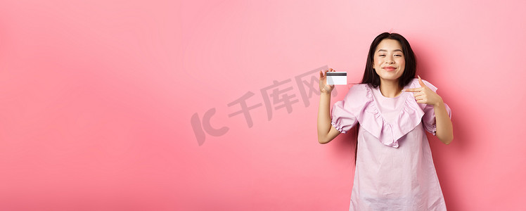 女性钱摄影照片_开朗可爱的亚洲女性满意地微笑着，展示塑料信用卡，穿着粉色背景的裙子