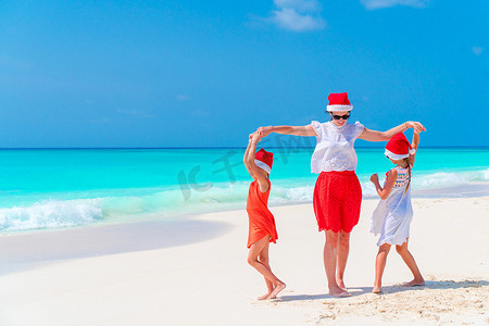 快乐美丽的母亲和孩子们在热带海滩庆祝圣诞假期时戴着红色圣诞帽