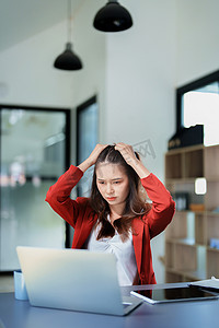 一位亚洲年轻女性的画像，她在清晨使用办公桌上的手机、财务文件和笔记本电脑时表情严肃