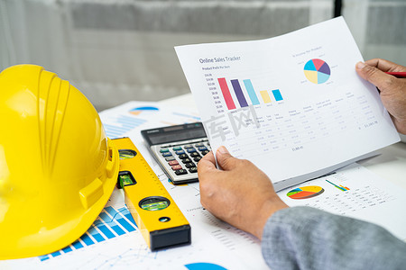 具有建筑房屋计划项目蓝图、图表和黄色头盔以及工程施工工具的亚洲工程师。