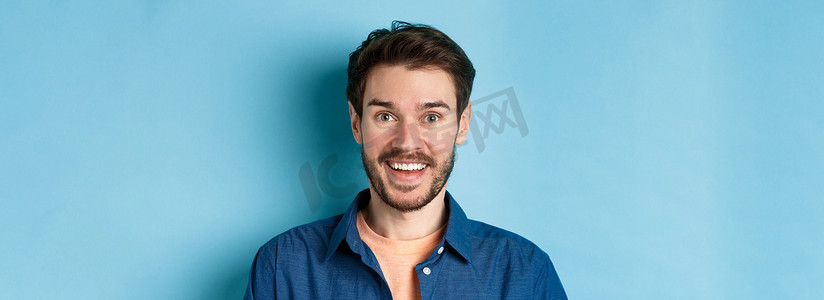 对镜头笑摄影照片_特写：留着胡子的惊讶帅哥看起来很有趣，对着镜头笑得很开心，站在蓝色背景上