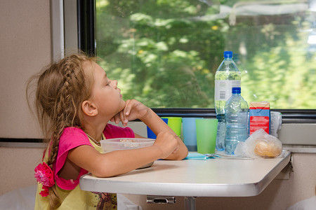 火车上一个六岁的女孩坐在一张桌子旁，食物放在车厢二等车厢的较低位置，若有所思地看着窗外