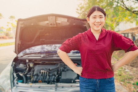 打开引擎盖摄影照片_肖像快乐的女人站在破损的事故车前，打开引擎盖，微笑着在路边等待汽车保险，不用担心。