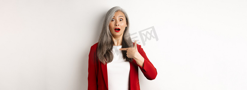 灰白背景摄影照片_惊讶的亚洲女性，头发灰白，指着自己，困惑地喘着粗气，站在白色背景上