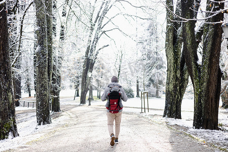 运动型父亲背着他的婴儿儿子穿着冬季连身衣和帽子在背包背袋中在冬天在城市公园散步的后视图