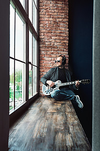 男歌手坐在窗台上，戴着耳机，吉他在家庭录音室录制曲目
