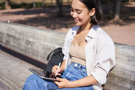 年轻的亚洲创意女孩用笔在平板电脑上画画，坐在公园的户外，画画
