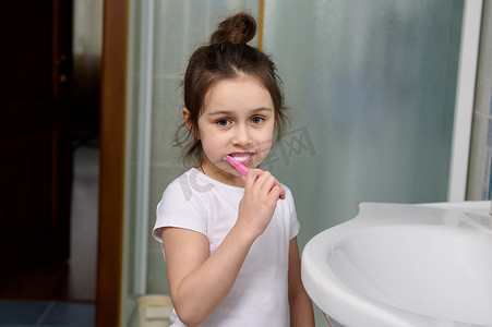 浴室儿童摄影照片_美丽的学龄前儿童小女孩一边刷牙一边对着镜头微笑，站在家庭浴室的水槽旁。