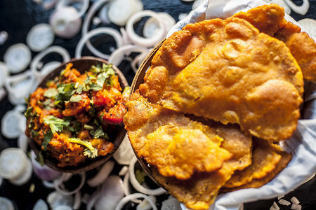 土豆地摄影照片_Batate ki sukhi bhaji 或土豆 sabji 在粘土碗中的特写镜头，以及一些干燥的普通油炸 puri 和一些黑色表面上的切洋葱。