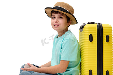 身穿蓝色 T 恤和带手提箱和登机牌的草帽的十几岁男孩看着相机，在白色背景中突显