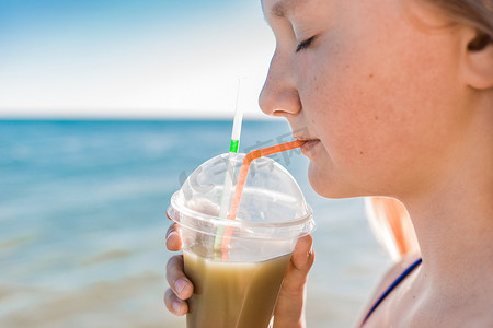 一个留着欧洲外表的金发的年轻女孩，在海滩的背景下，一个少年手里拿着并喝着一杯彩色的冷无酒精鸡尾酒