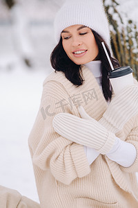 杯子里的咖啡摄影照片_一个穿着米色开衫和白帽子的漂亮女孩在湖边的雪冬森林里喝茶