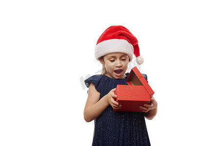 红色大气礼盒摄影照片_戴着圣诞帽的小女孩惊讶地表达兴奋，同时打开一个装有可爱圣诞礼物的红色礼盒