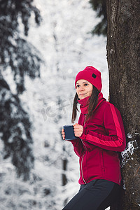 雪林中身穿红色夹克的美女靠在树上的画像，手里拿着一个可重复使用的金属杯，里面装着热茶