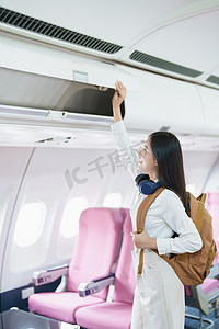 美女手提行李摄影照片_年轻的亚洲美女乘飞机旅行，乘客戴着耳机把手提行李放在飞机座位上方的储物柜里