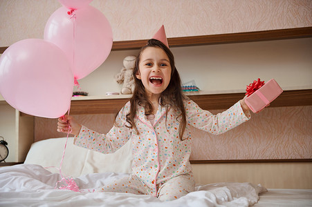 戴着派对帽的快乐生日女孩，拿着一堆粉色气球和礼盒，坐在床上，对着镜头微笑