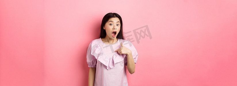 惊讶的亚洲女孩喘着粗气想知道，指着自己，被选中或挑选，站在粉红色背景下的裙子