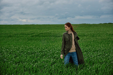 一个穿着外套的女人站在田野中间，在多云的天气里，在强风中侧身对着镜头