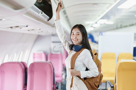 手提行李摄影照片_年轻的亚洲美女乘飞机旅行，乘客戴着耳机把手提行李放在飞机座位上方的储物柜里