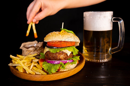 三明治摄影照片_美味的汉堡汉堡三明治配薯条和番茄酱在木盘上一杯冰镇啤酒泡沫快餐黑色背景。