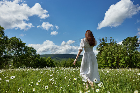一个长着红色长发的女人背对着镜头站着，身着浅色连衣裙站在蓝天映衬下的雏菊花田里，望着远方