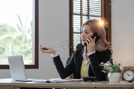 年轻的亚洲女商人坐在笔记本电脑前，愉快地与客户通电话，解释办公室里的细节。