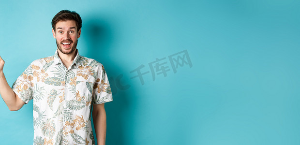 惊讶的快乐游客用手指指着空地，展示酷炫的促销，站在蓝色背景中的夏威夷衬衫
