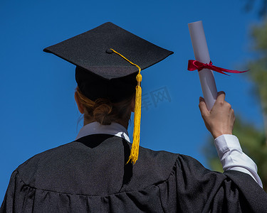 身着毕业礼服的白种女人背影拿着文凭，映衬着蓝天。