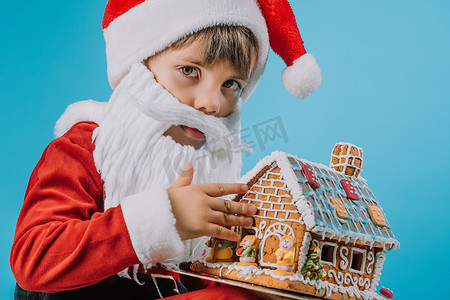 穿着圣诞老人服装的可爱小孩坐在蓝色的姜饼屋里
