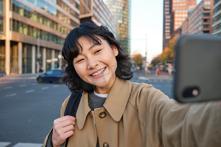 亚洲年轻女性在市中心大楼前自拍的肖像，游客在观光时拍照，对着智能手机相机微笑