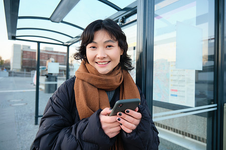 可爱的小女孩，学生在公共汽车站等车，在应用追踪器上寻找她的公共交通工具，拿着智能手机，站在路边的街道上