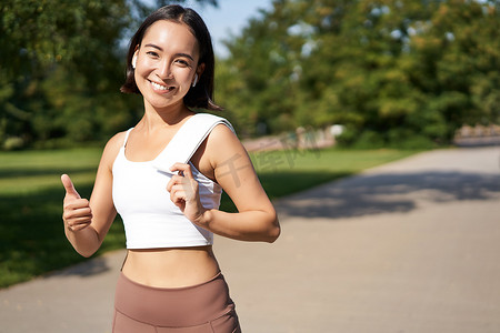 微笑的健身女孩竖起大拇指，在公园锻炼身体后拿着毛巾站着，慢跑，看起来很开心