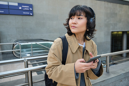亚洲女性的肖像，戴耳机的学生，拿着智能手机站在车站的街道上，等人，乘坐公共交通工具上下班