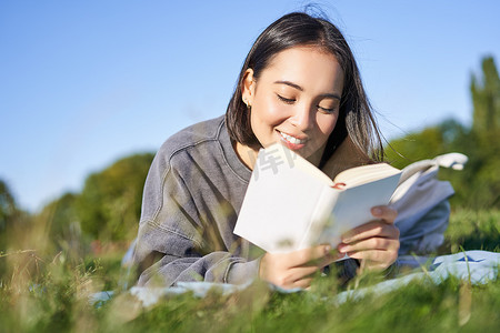 读书器摄影照片_可爱的韩国女孩的画像，在公园里躺在草地上读书，手里拿着最喜欢的书放松，开心地笑着