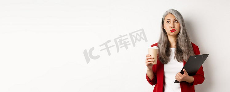 疲惫背景摄影照片_疲惫的亚洲女上班族拿着剪贴板和纸杯，喝着咖啡，脸色疲惫地呼气，站在白色背景上